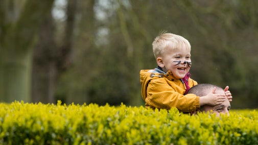 A young child in a yellow coat sits on his fathers shoulders, laughing. Only the top of the father's head is visible behind the top of a hedge in a maze at Belton.